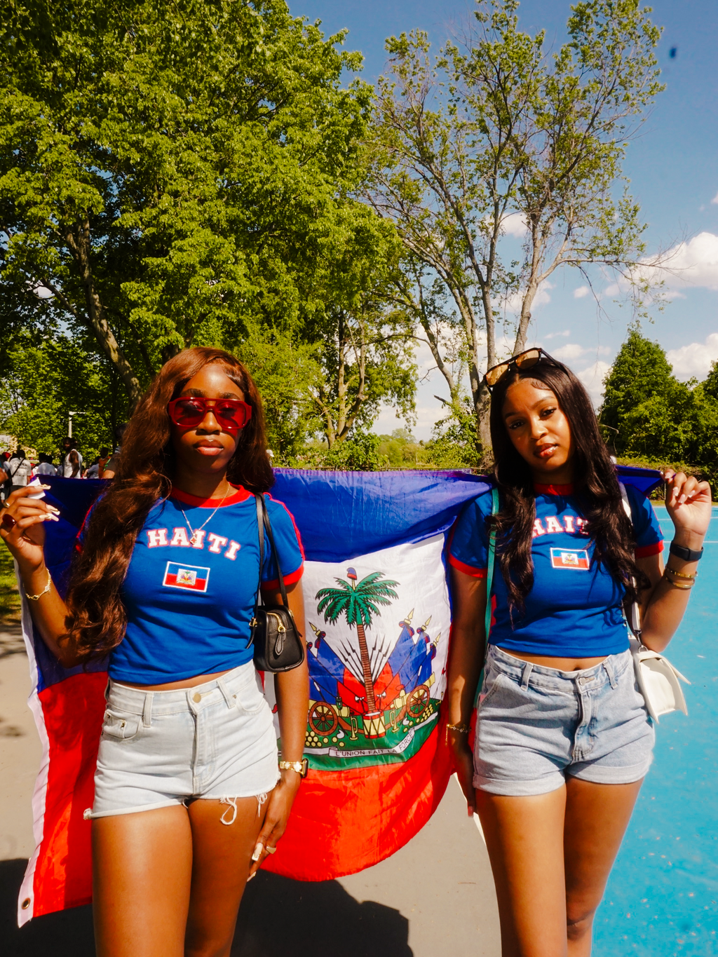 Two women holding a Haitian flag outdoors with trees and blue sky in the background.