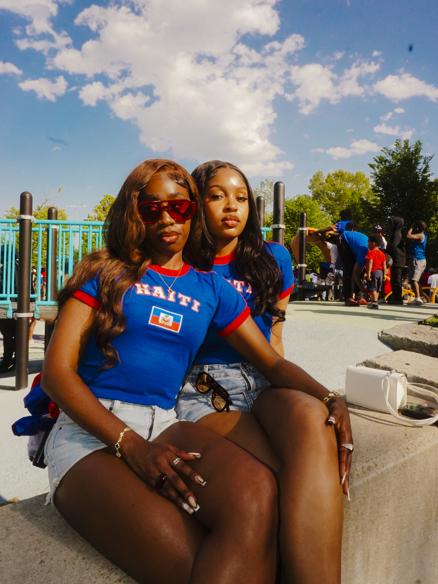 Two women sitting on a concrete ledge wearing blue shirts with 'Haiti' printed on them, against a blue sky with clouds.