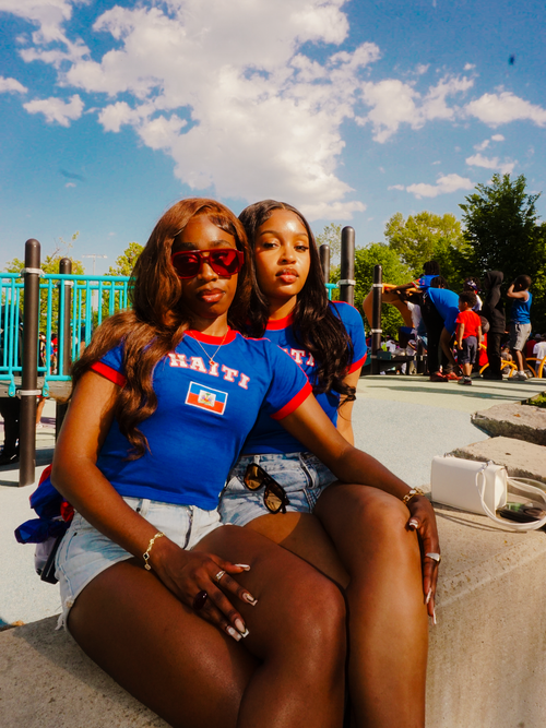 Two women sitting on a concrete ledge wearing blue shirts with 'Haiti' printed on them, against a blue sky with clouds.