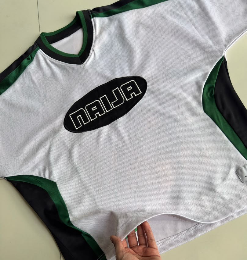 A white athletic jersey with green and black trim, featuring a black oval chest logo with the word 'NAIJA' in white. A hand is holding the hem of the shirt, which is laid flat on a light beige tiled floor.