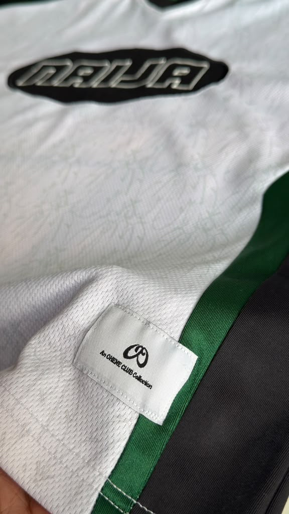 Close-up of a white athletic jersey with a black and green 'Naila' logo on the chest, a green side stripe, and a sewn-in size/brand label reading 'AN OHENE Club Collection'.