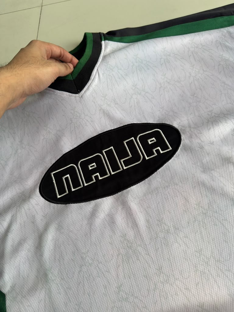 A close-up of a white sports jersey with a black oval patch reading 'NAIJA' in white letters, held at the collar by a hand. The jersey has green and black trim at the collar and shoulders.