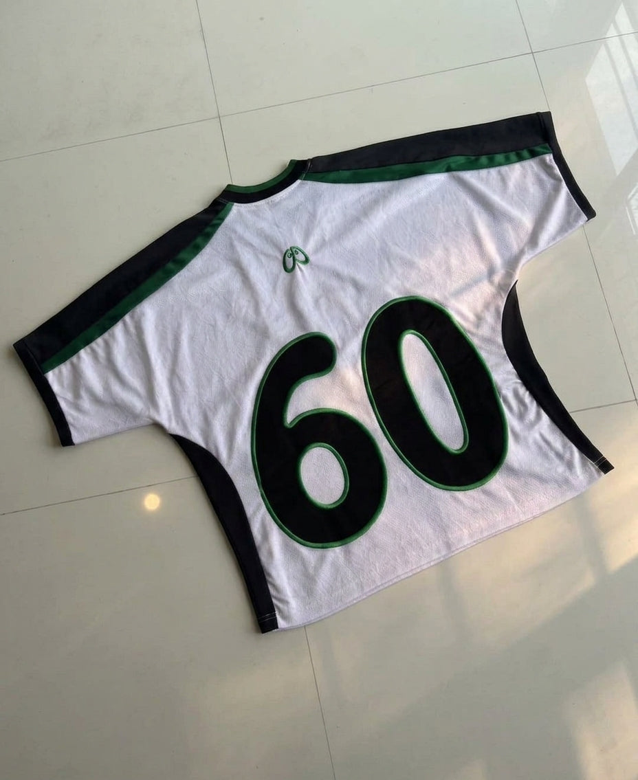 A white short-sleeve athletic jersey laid flat on a light tiled floor, featuring black shoulder and side panels with green trim, and large black number 60 outlined in green on the back.